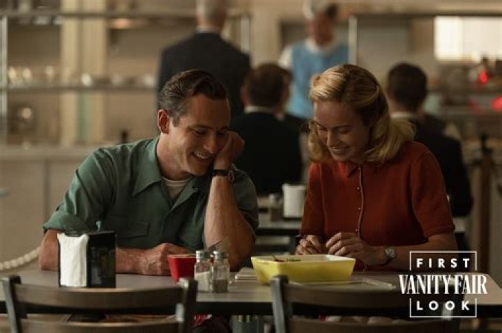 Lewis Pullman and Brie Larson as Calvin and Elizabeth Zott sitting next to each other at a cafeteria table smiling in Lessons in Chemistry on Apple TV